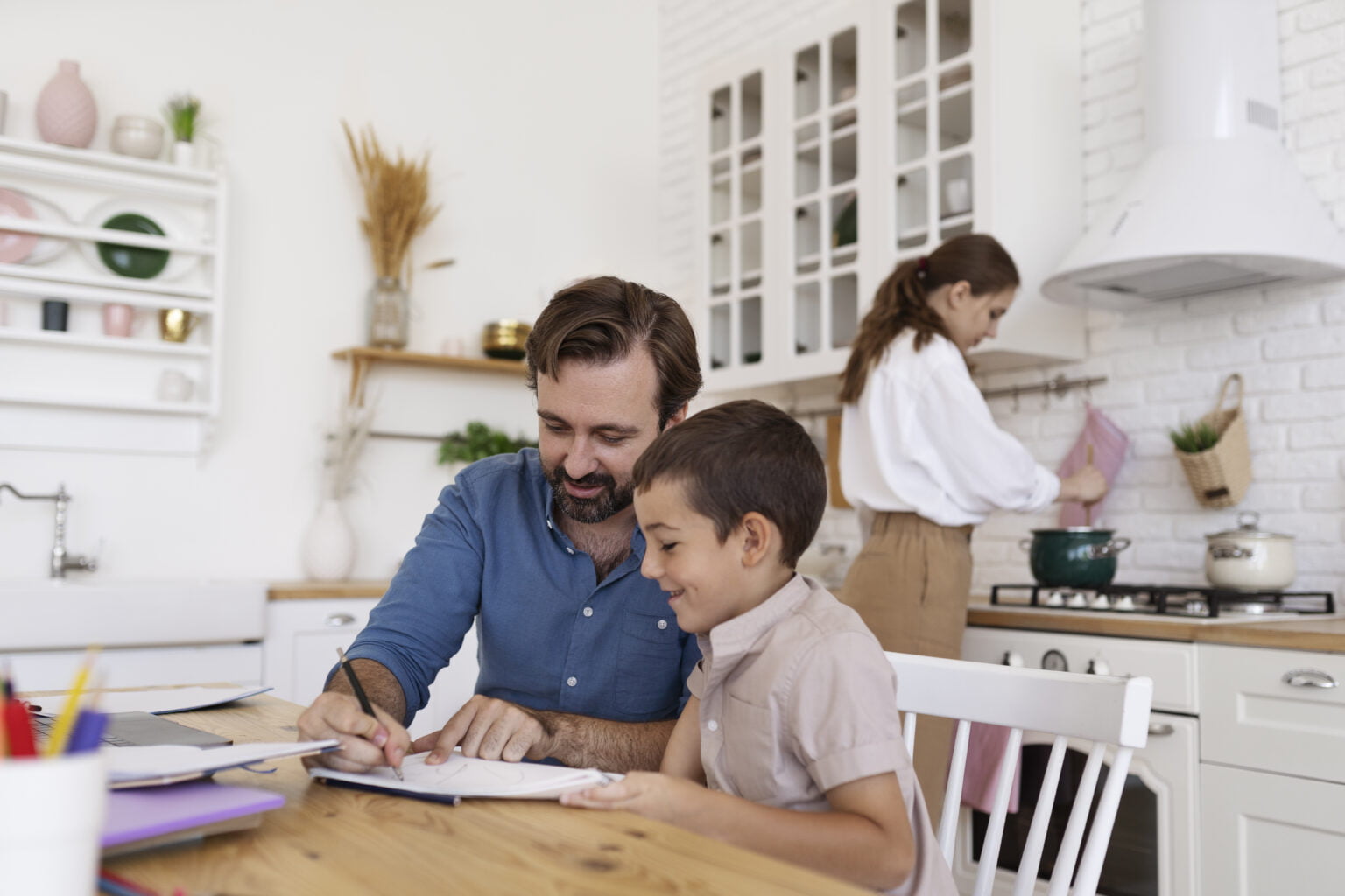 pai e filho sentados à mesa, enquanto pai ensina lição de casa ao filho, e mãe cozinha no fundo