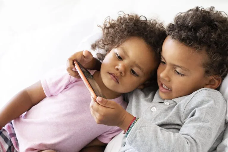 Duas crianças pequenas, provavelmente gêmeas do mesmo sexo, estão deitadas em uma cama, observando atentamente algo em um smartphone. A criança da esquerda veste uma camisa rosa; a da direita ostenta uma camisa cinza. Ambas têm cabelos cacheados e estão focadas intensamente na tela, exibindo sua dinâmica única de gêmeas.