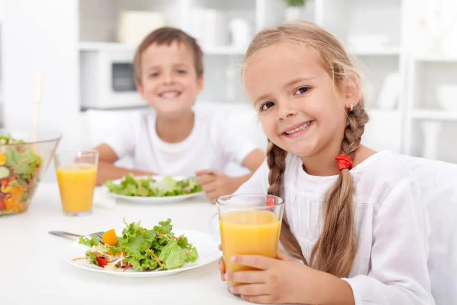 Crianças sorridentes tomando suco natural e comendo salada, representando alimentação saudável na infância.
