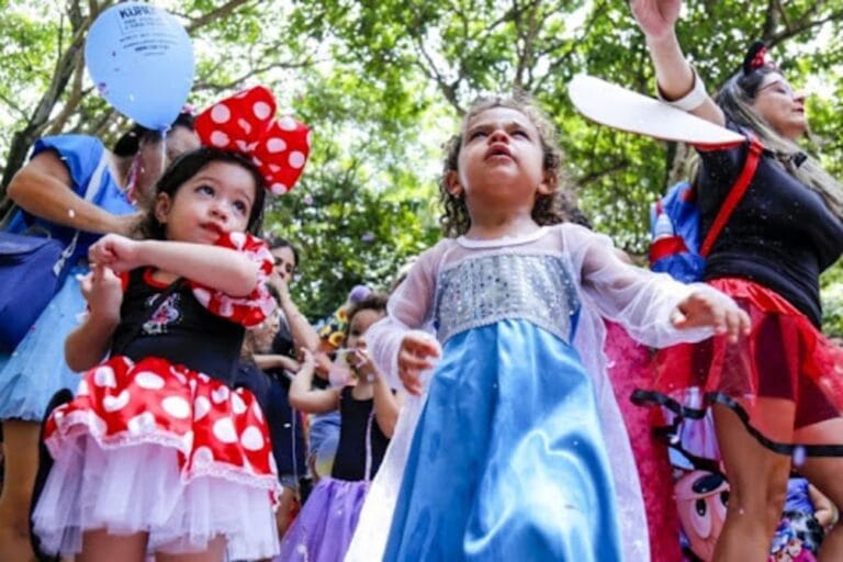 Crianças fantasiadas de Minnie e princesa curtindo o Carnaval em um bloco infantil, cercadas por foliões em clima de festa.