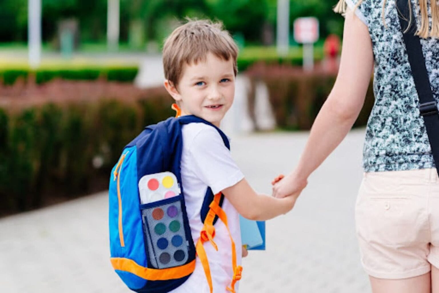 Menino com mochila azul sorrindo enquanto segura a mão de um adulto, representando o retorno seguro às aulas na educação infantil.